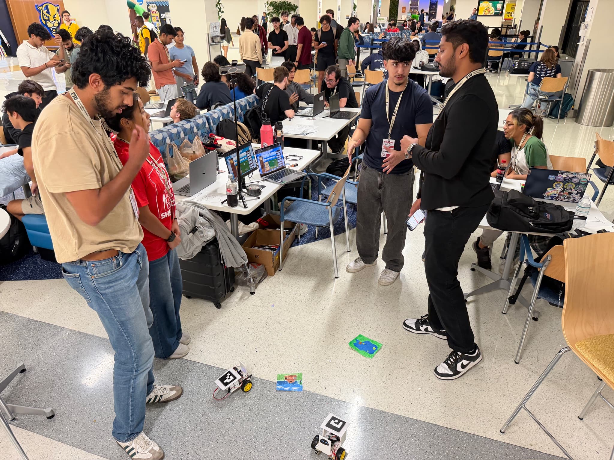 A robotics demonstration at ShellHacks, with two small robots on the floor and laptops running nearby.